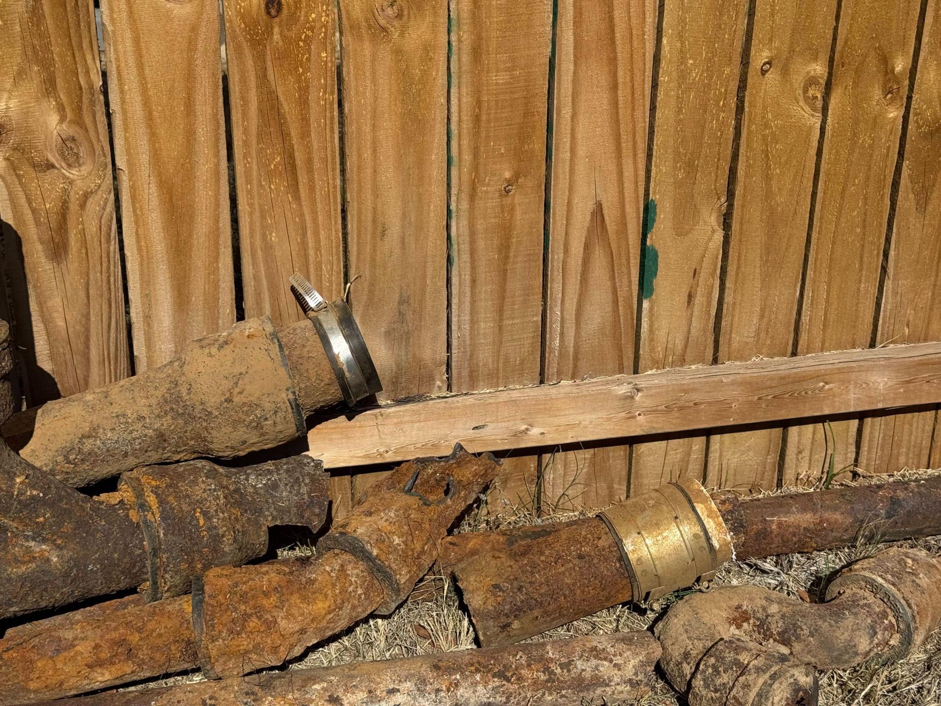 Rusty pipes and fittings piled against a wooden fence.