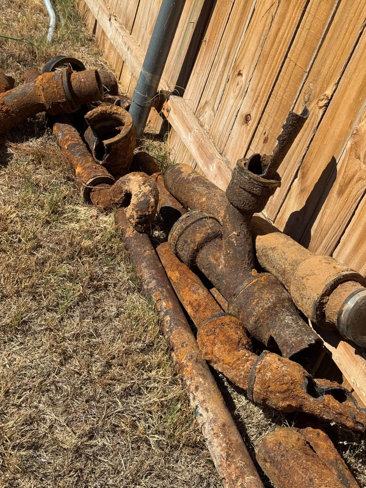 Rusty metal pipes and fittings piled on dry grass next to a wooden fence.