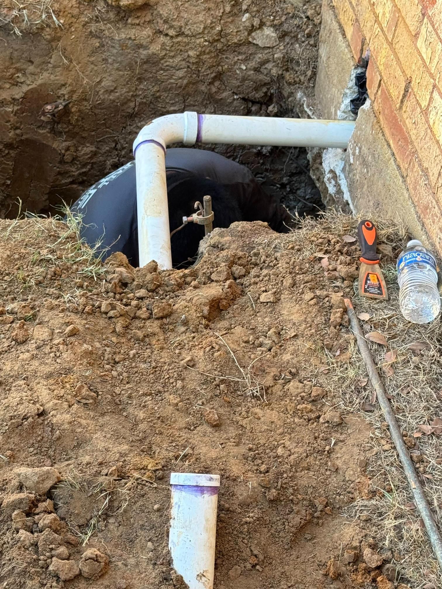 Excavation revealing buried pipe and tank near a brick wall; white PVC piping, dirt, and tools present.
