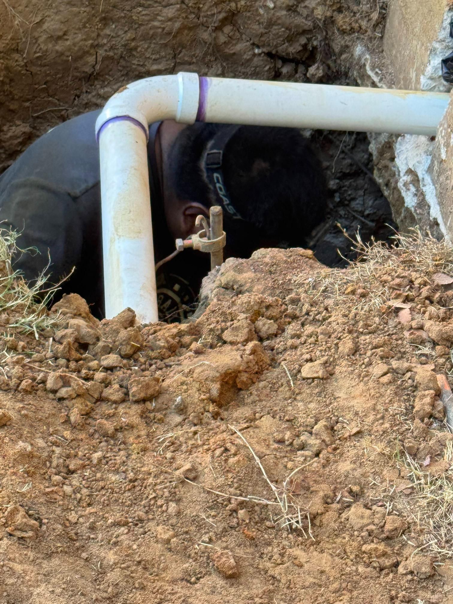 Person working on plumbing, buried in a hole with white PVC pipes.