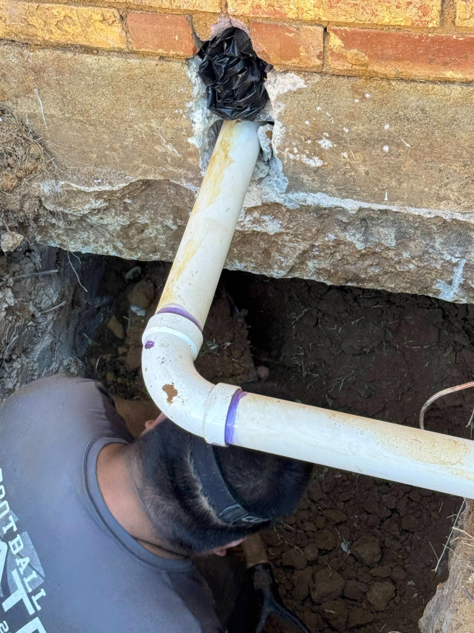 Person working on buried PVC pipe near a brick building. The pipe runs through a wall.