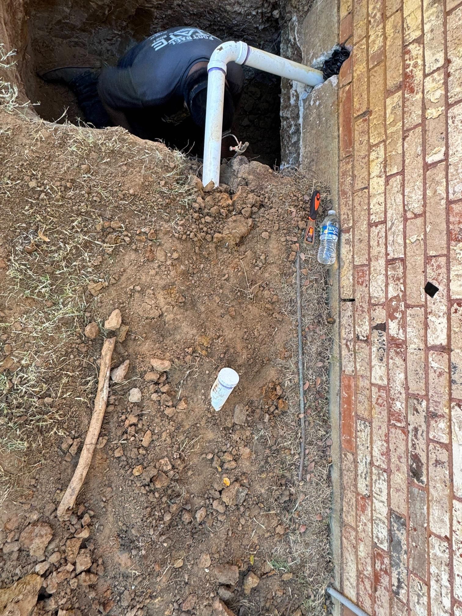 Person working on pipes in an excavated area near a brick wall. Tools and pipes are visible in the dirt.