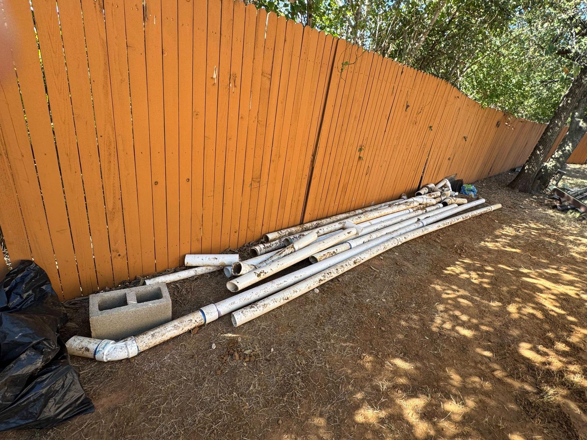 Pipes and cinder blocks rest on dirt next to an orange wooden fence in an outdoor setting.