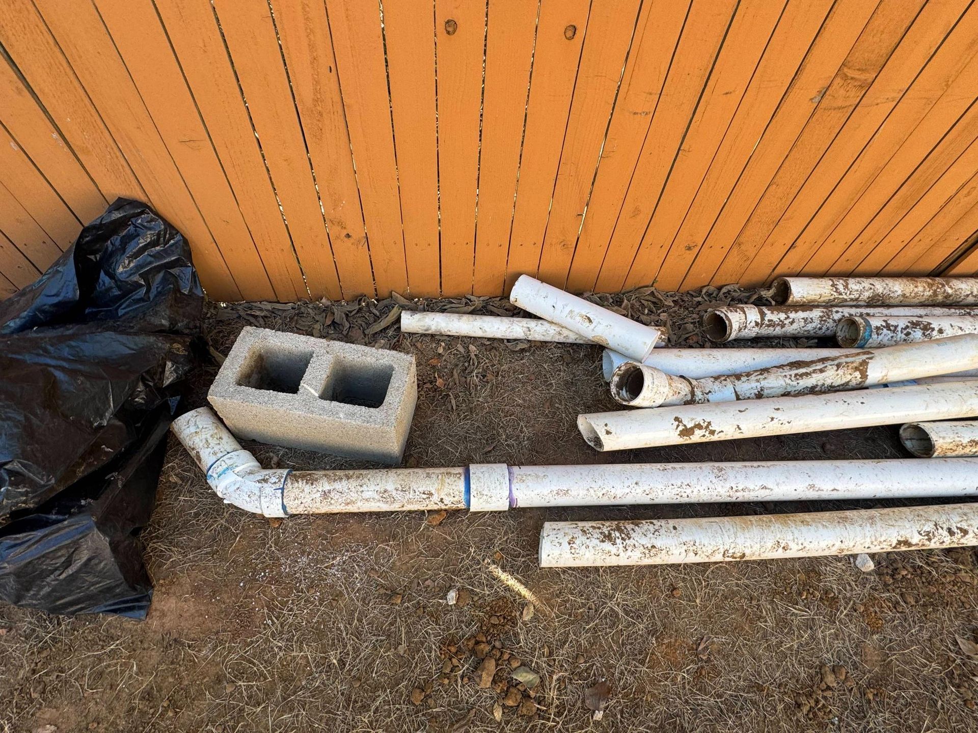 Pile of white pipes and cinder block on dirt ground near a wooden fence and black trash bag.