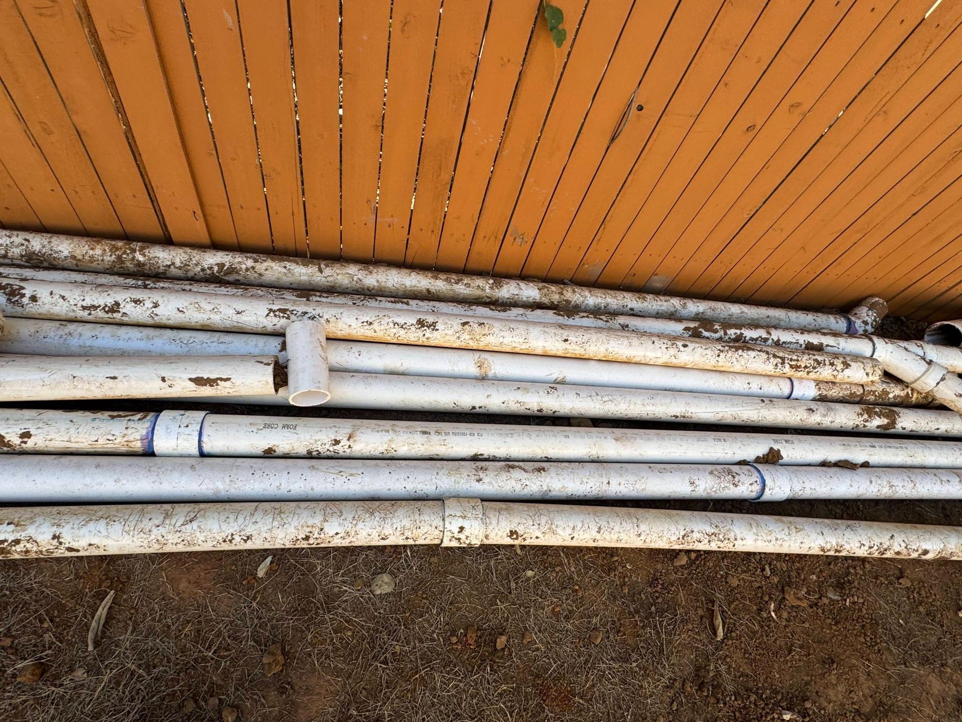 White PVC pipes on brown ground, leaning against a brown wooden fence.