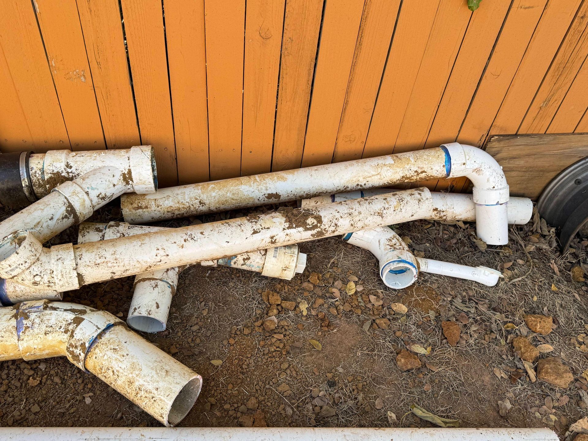 Pile of dirty white PVC pipes on brown dirt, against an orange wooden fence.