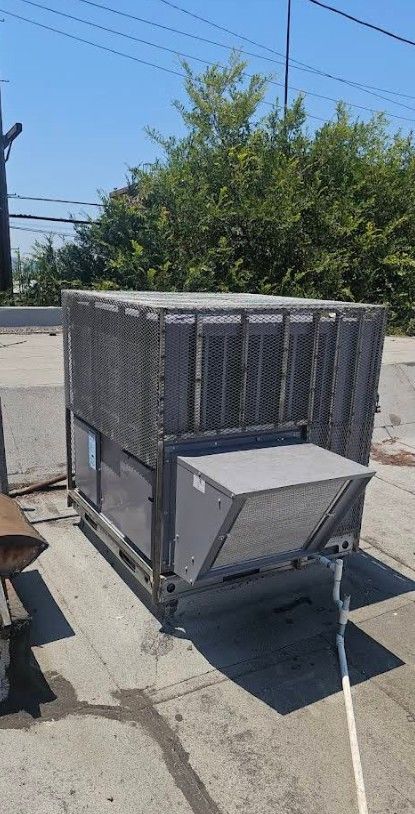 Gray industrial cooling unit on a rooftop, with a protective metal cage. Blue sky and green trees in the background.