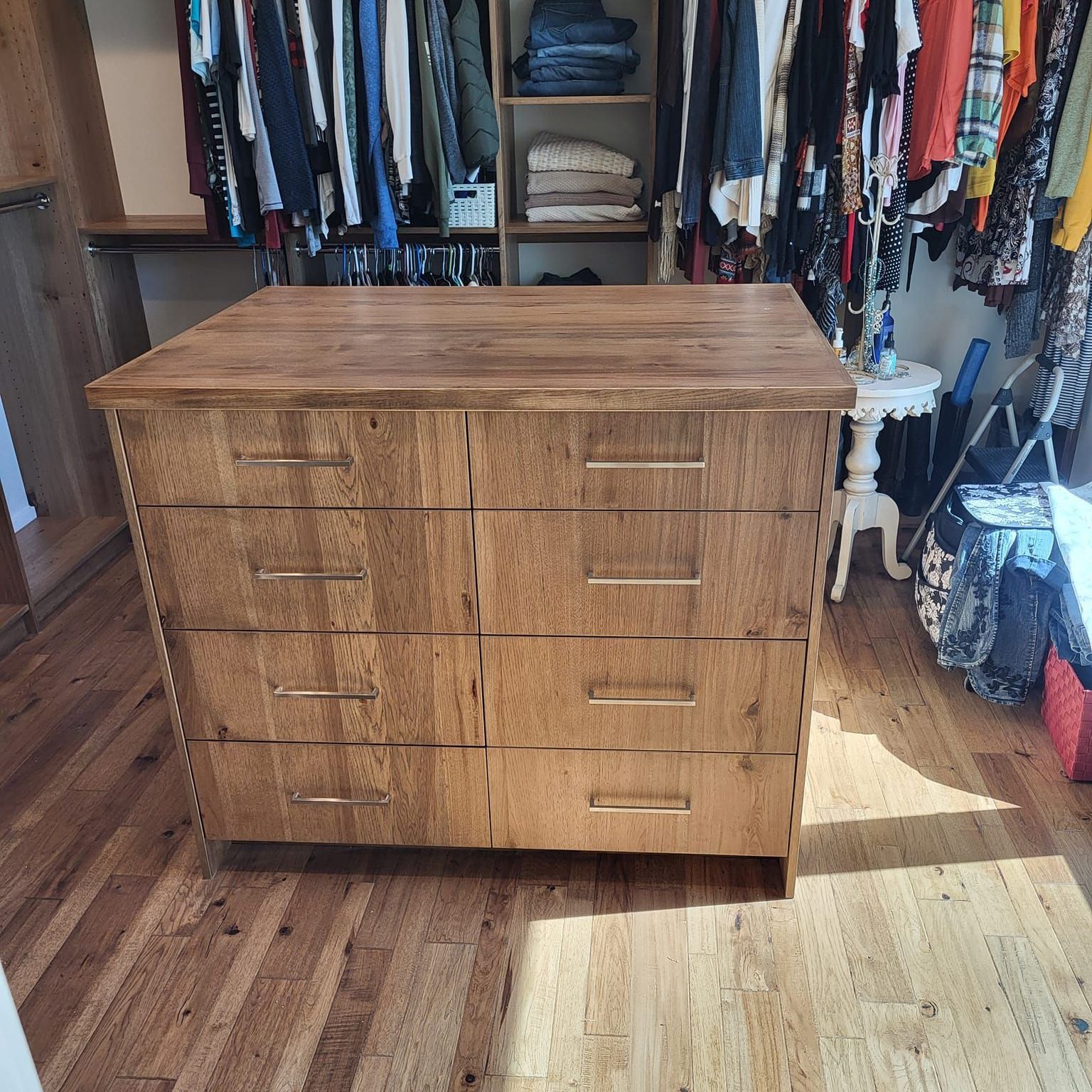 A wooden dresser is in a walk in closet filled with clothes.