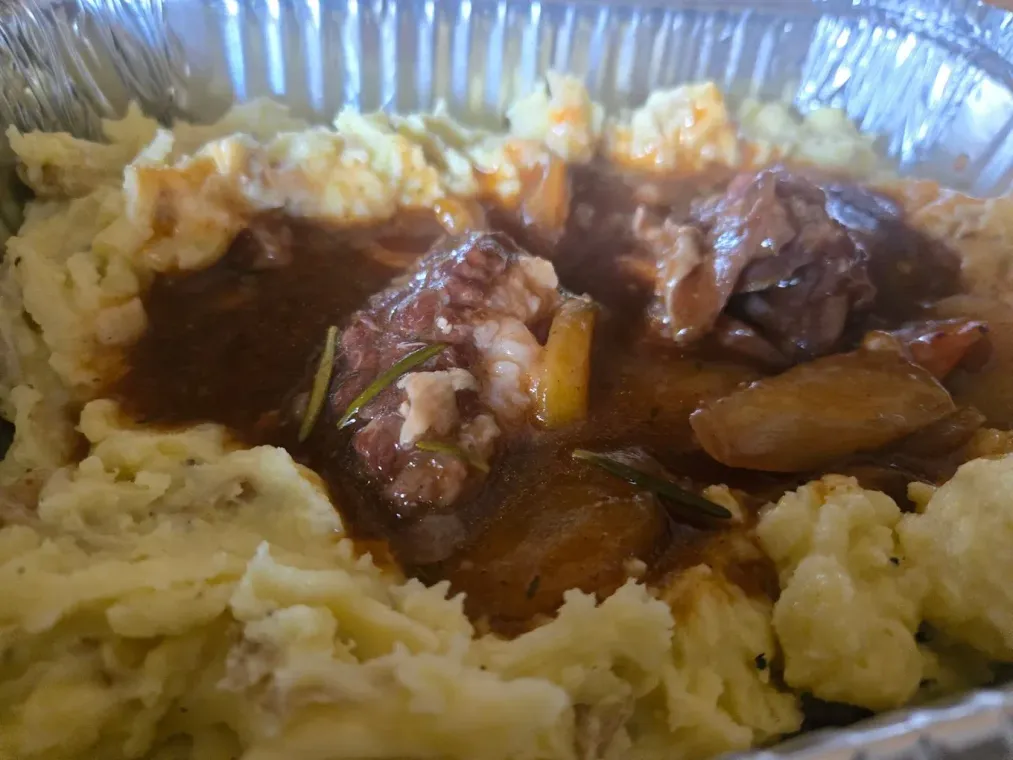 Mashed potatoes topped with braised meat in gravy, served in a foil container.