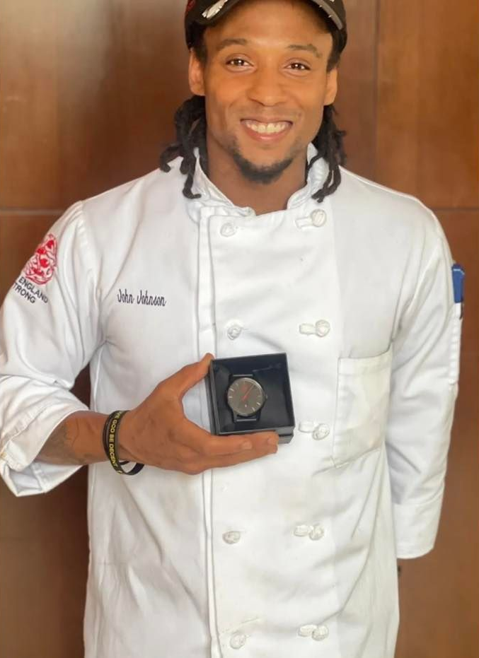 Chef holding a watch in a box, smiling. He wears a chef's coat and a baseball cap.