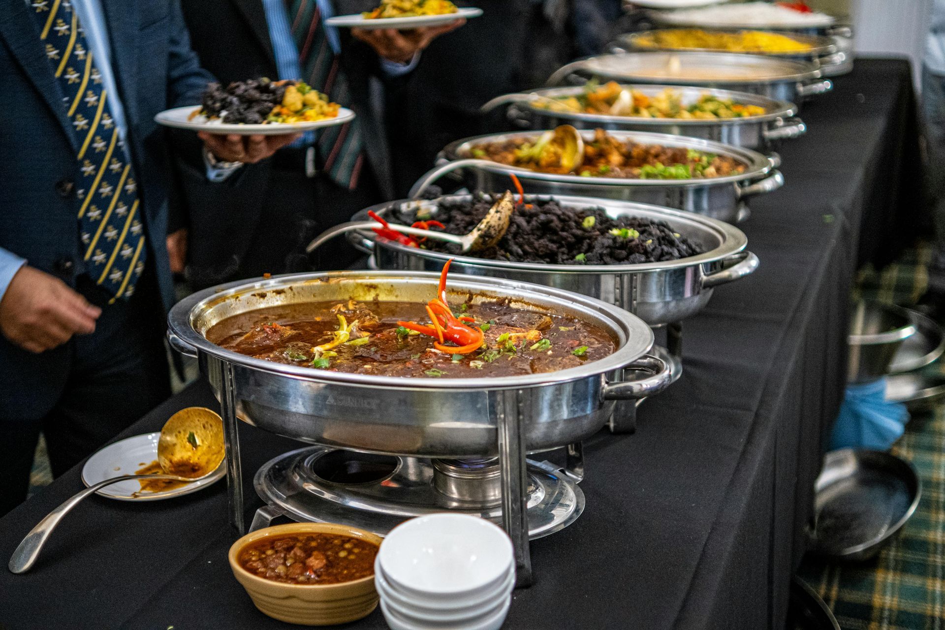 A buffet table with various dishes in chafing dishes, people holding plates, and serving utensils.