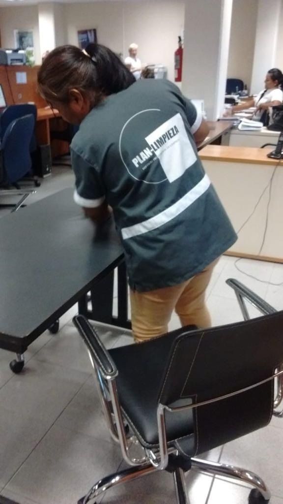 Una mujer está limpiando una mesa en una oficina mientras está parada junto a una silla.
