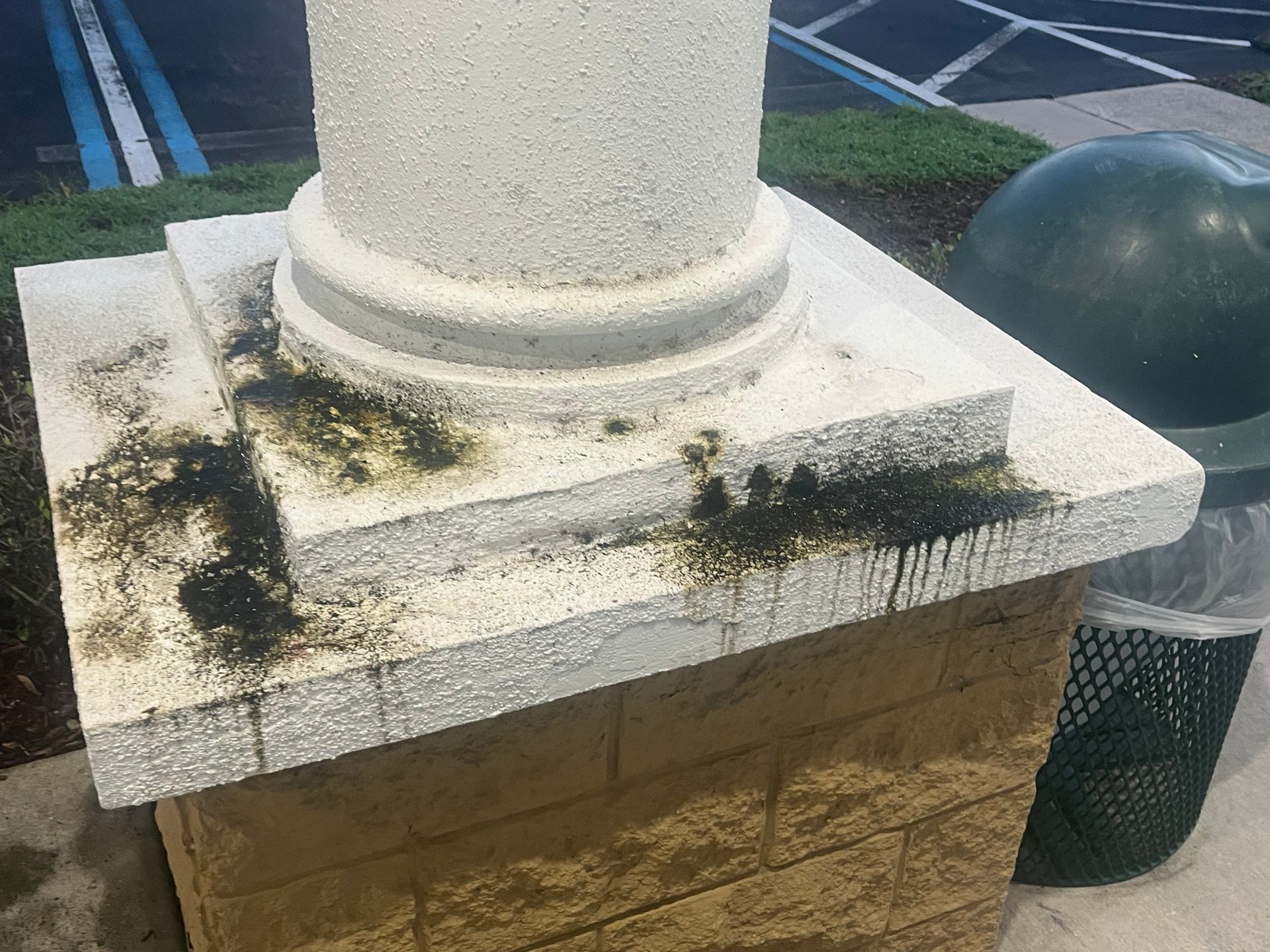 A white pillar with green mold and yellow brick base next to a green trash can.