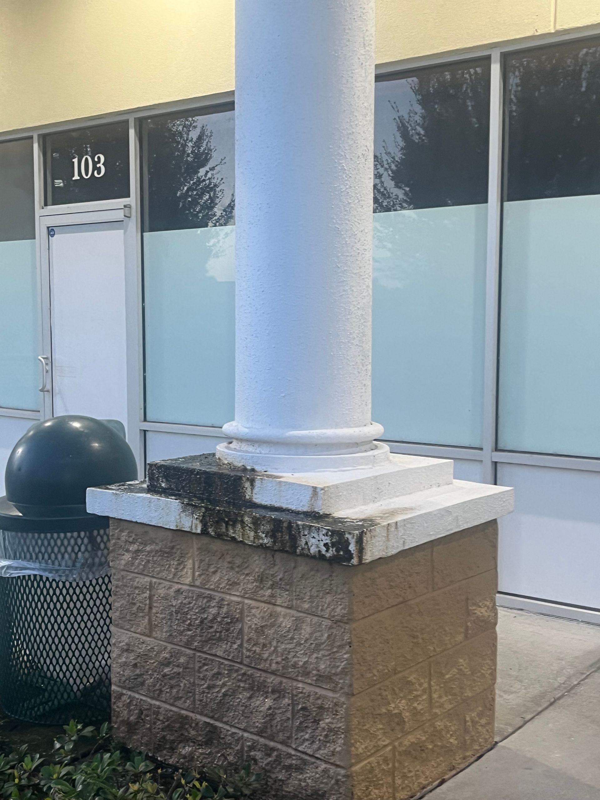 White column on a brick base, next to a trash can and building entrance.