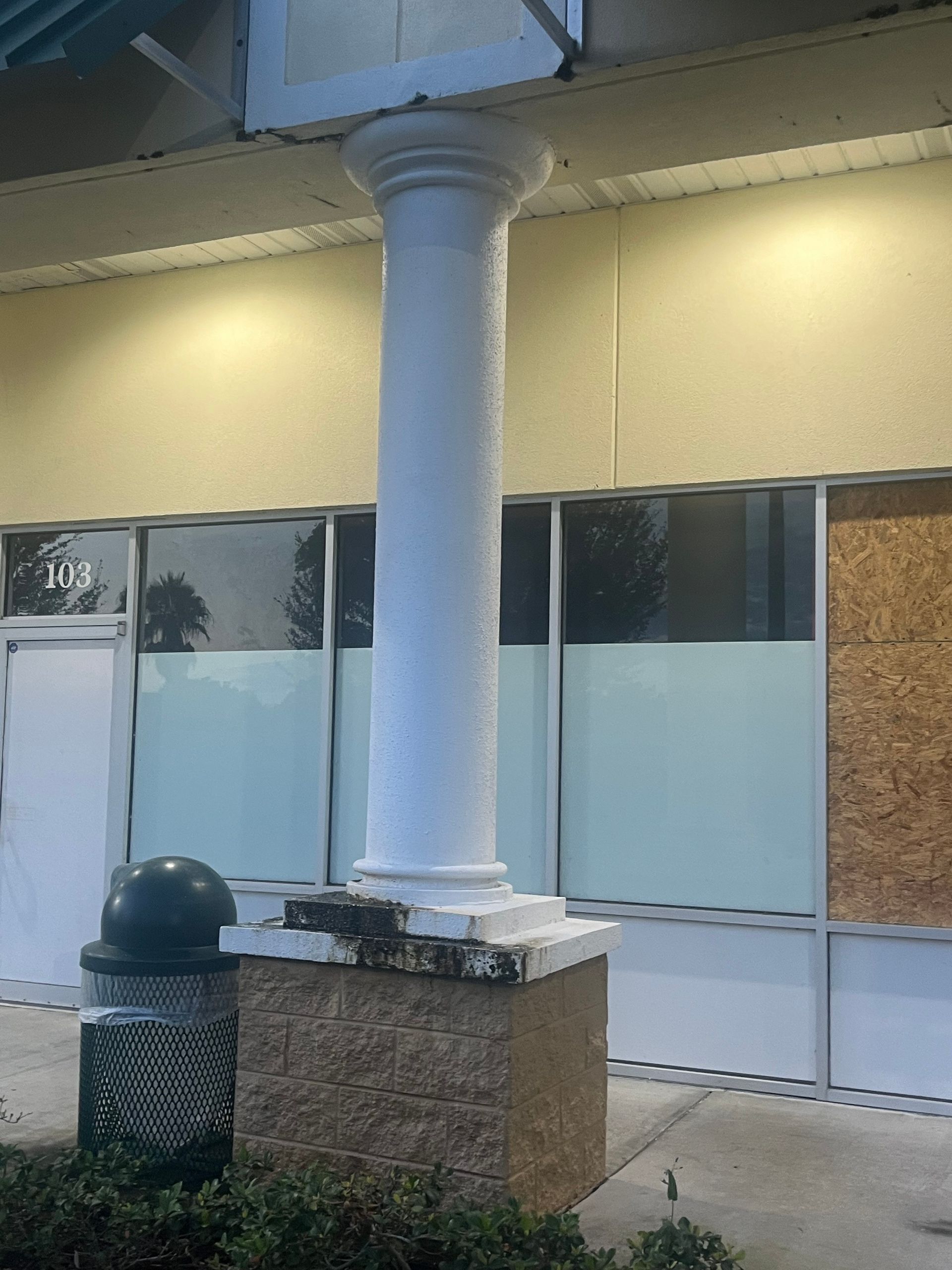 White column in front of a store with boarded-up windows and a trash can.