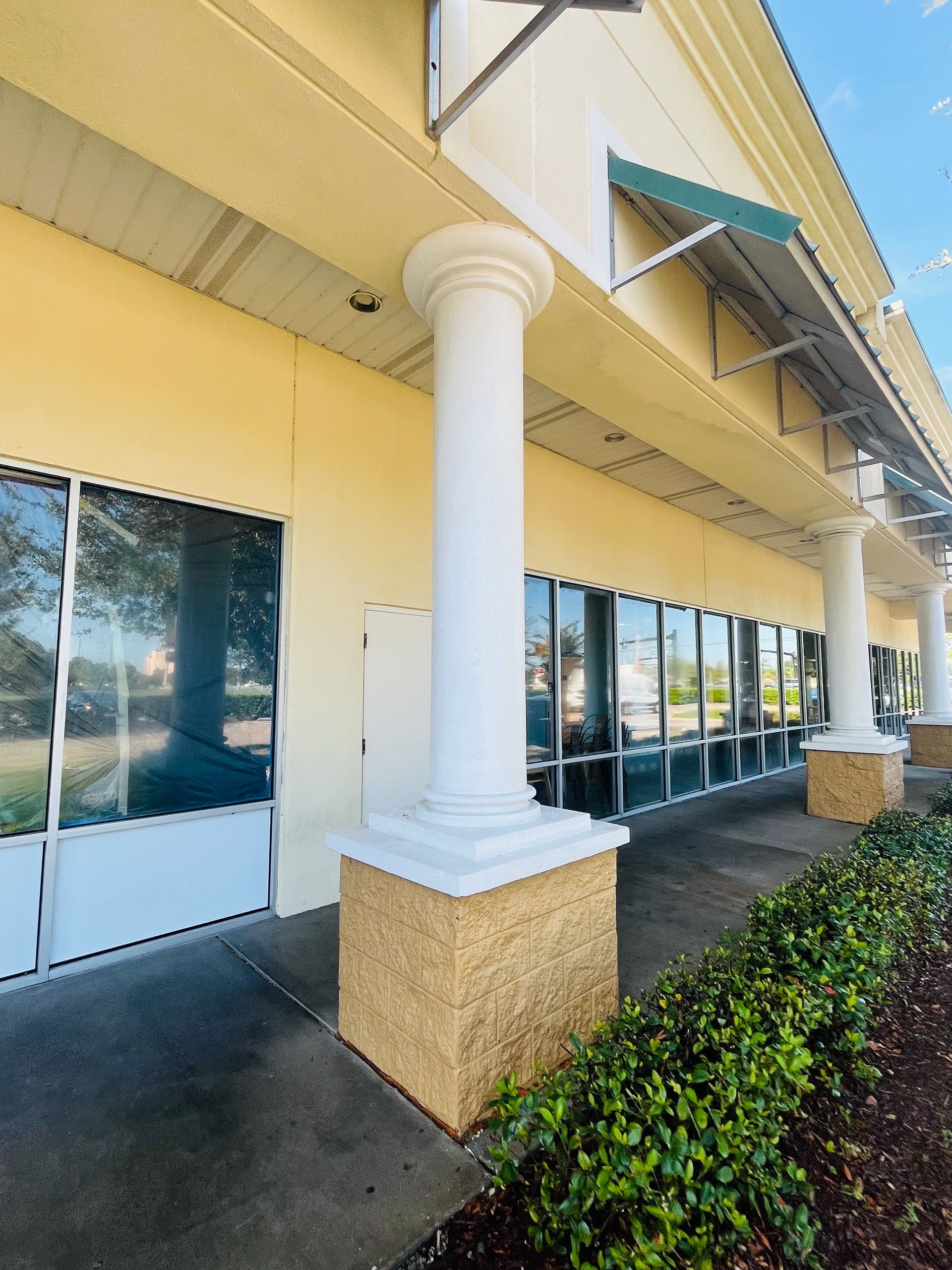 Exterior view of a building with columns and windows, with bushes in the foreground, and a blue sky.