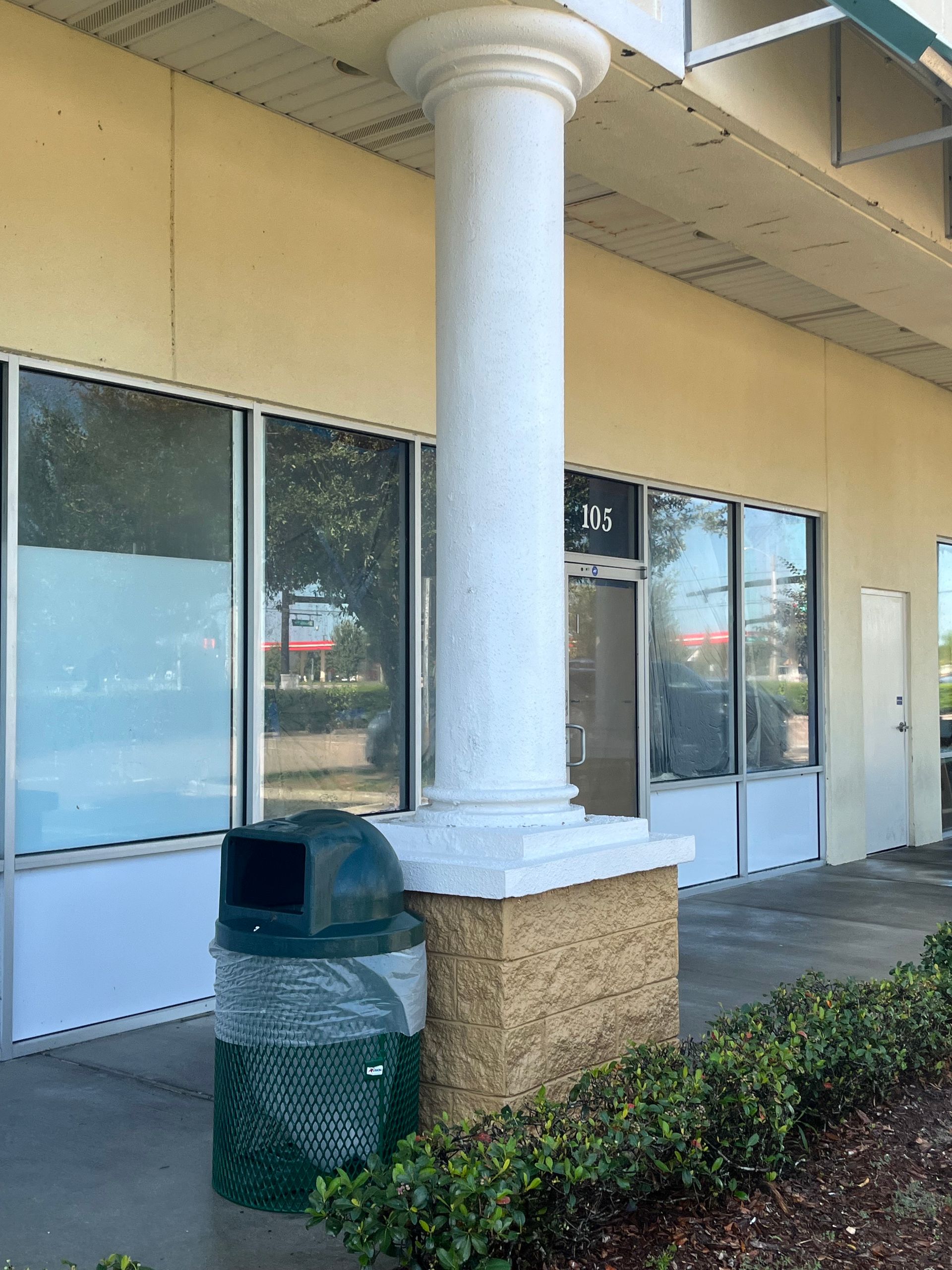 A trash can sits next to a white column in front of a beige commercial building with windows.