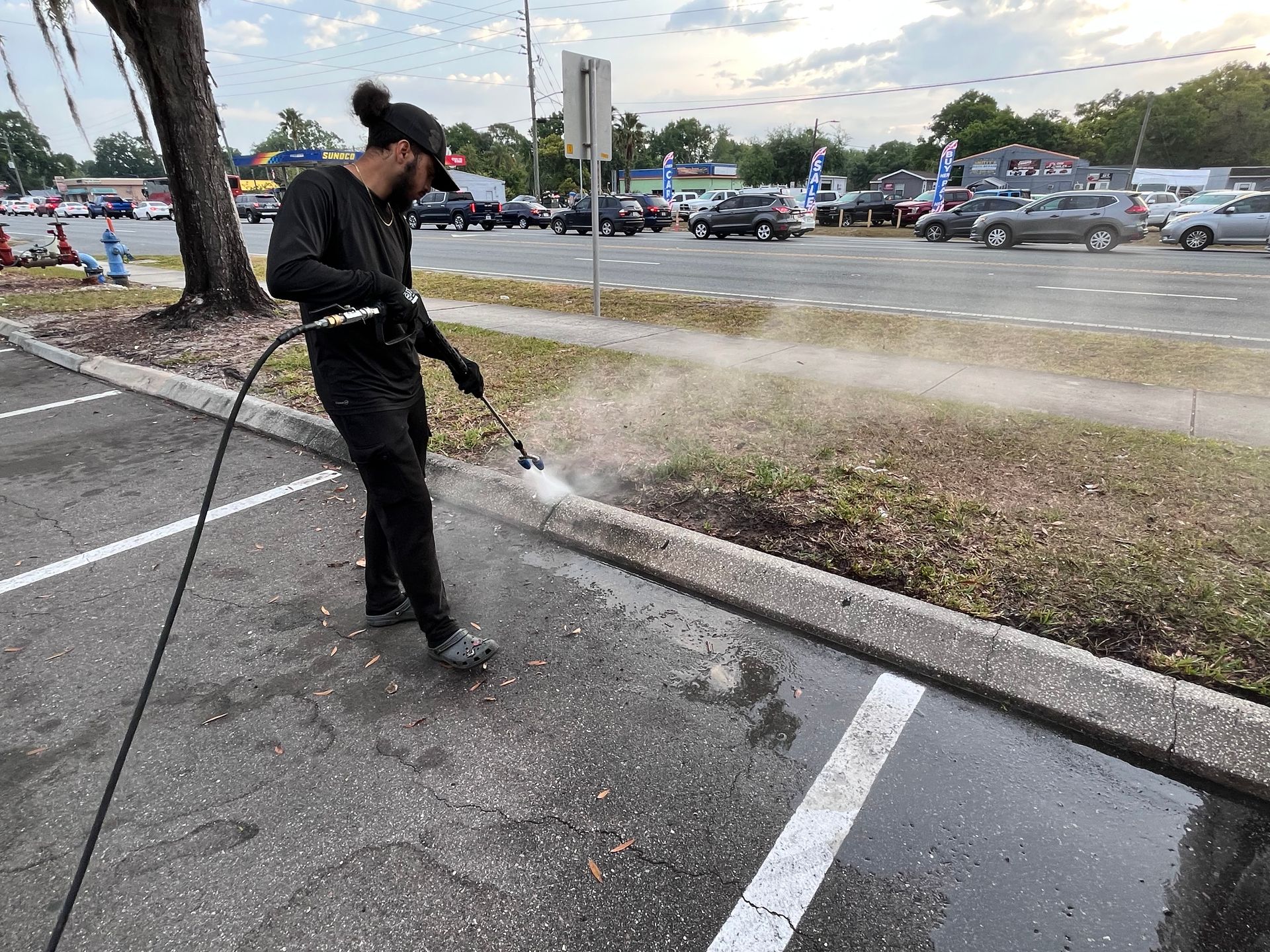Man in black clothes power washes a curb near a parking lot.