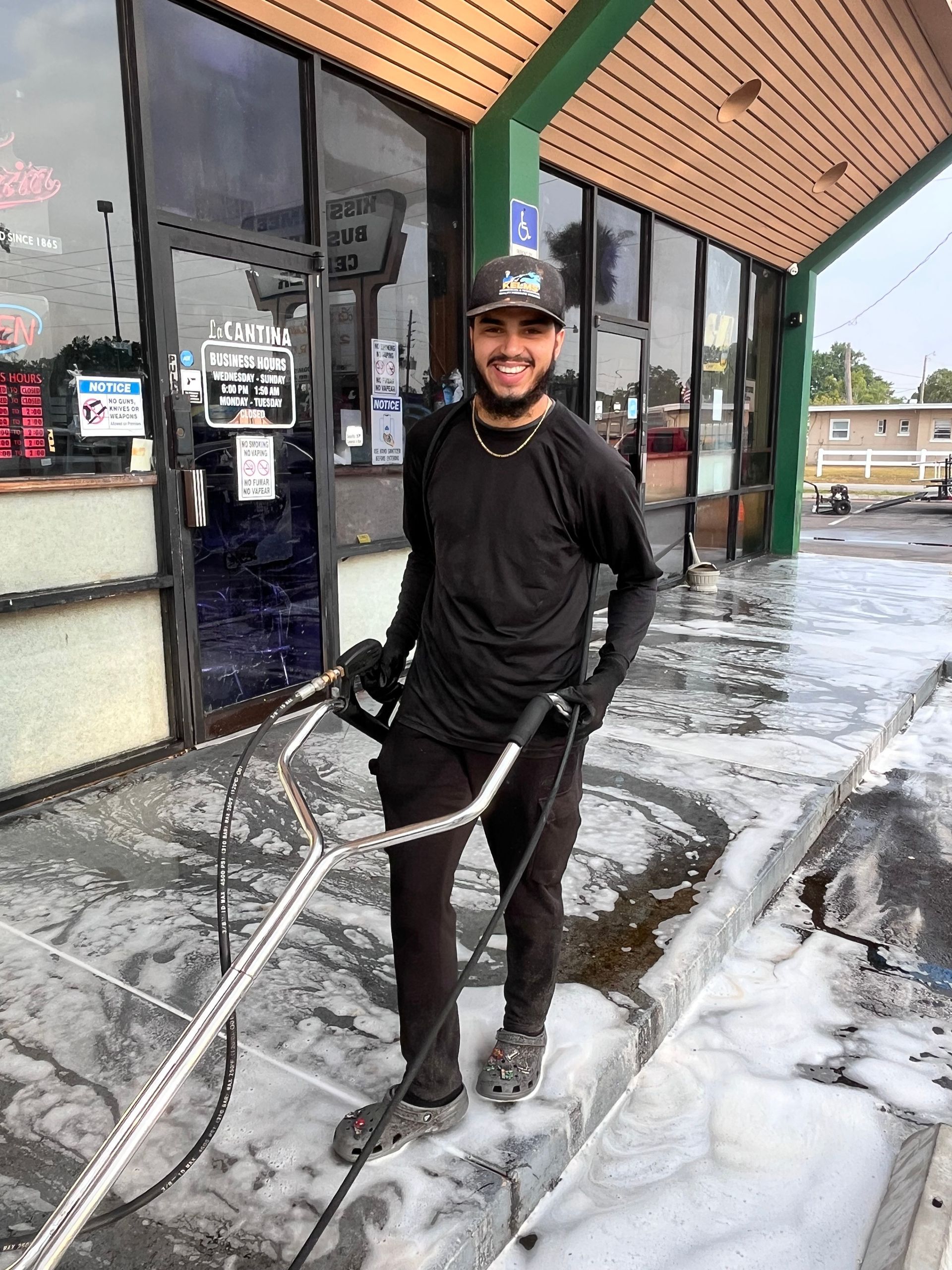 Man power washing a sidewalk in front of a building. He is smiling and wearing black clothes and a cap.