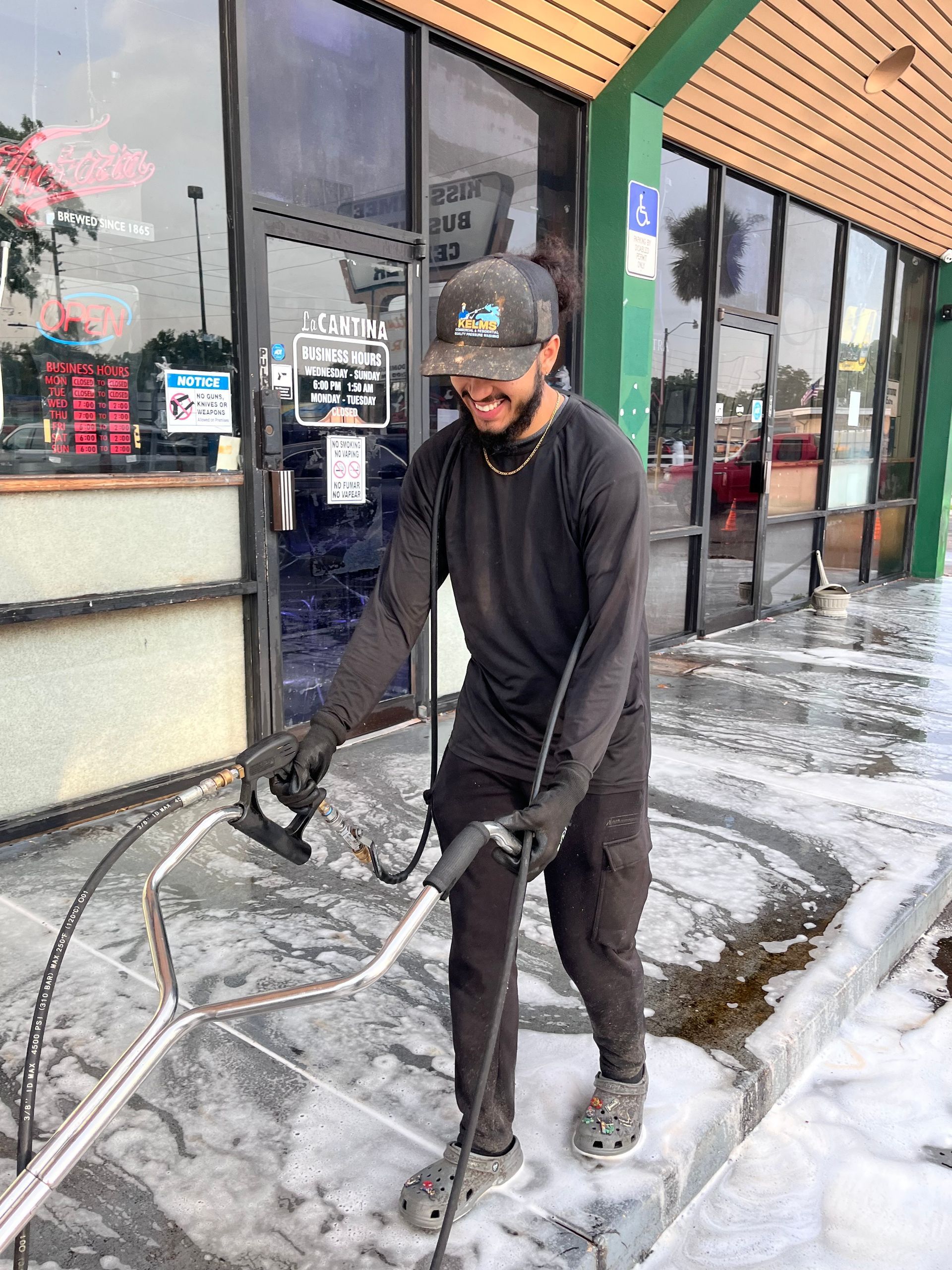 Man power washing a sidewalk in front of a business; he is smiling and wearing a hat.