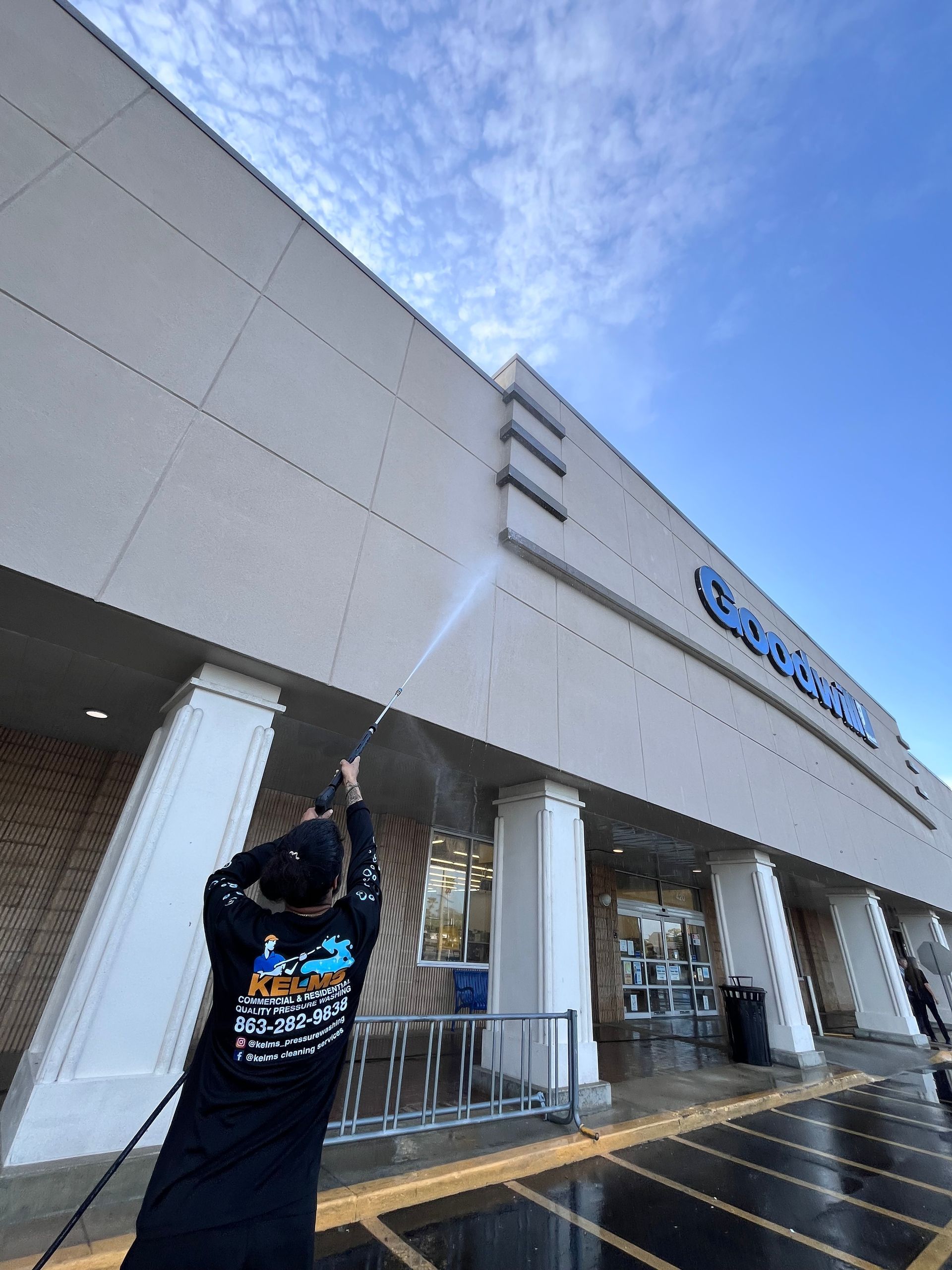 A person power washing the exterior of a Goodwill store under a blue sky.