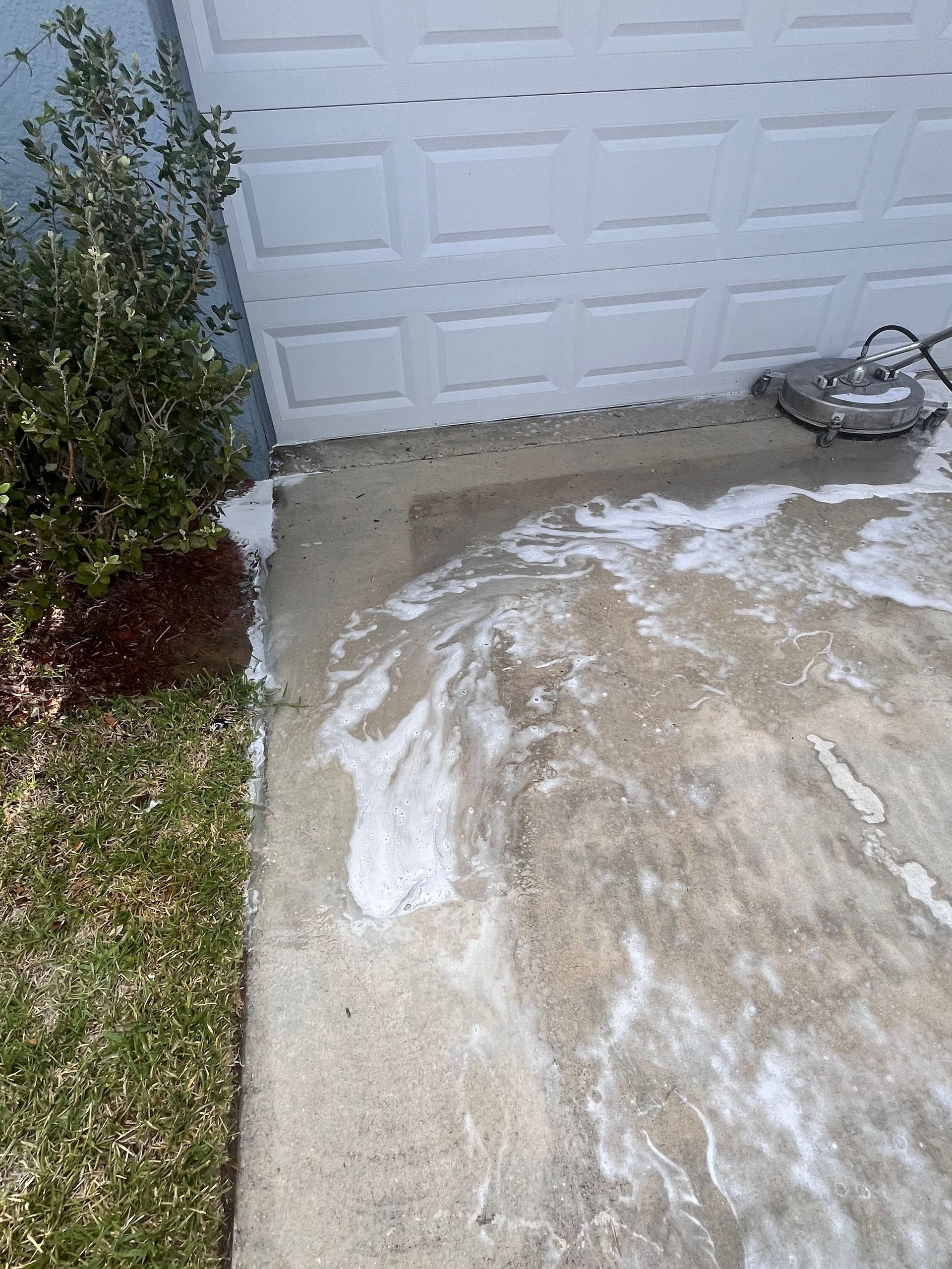 A person power washing a concrete driveway in front of a white garage door, with soap and cleaning residue.
