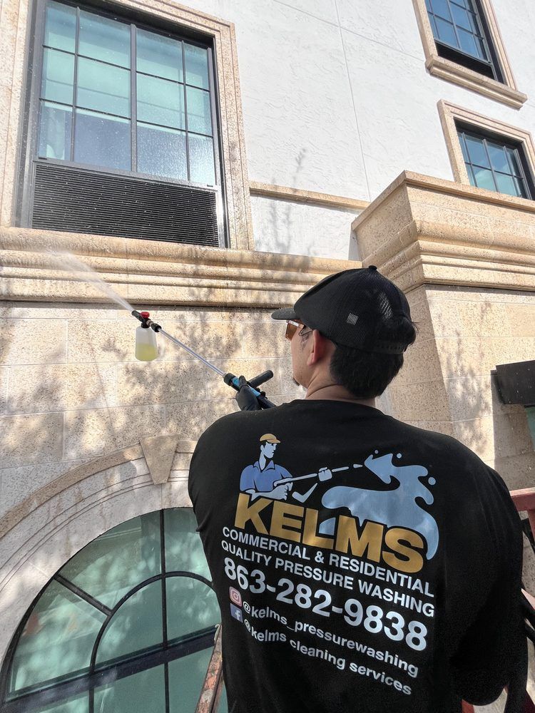 Man in a black Kelms pressure washing shirt spraying the side of a building. The building is white with dark-framed windows.