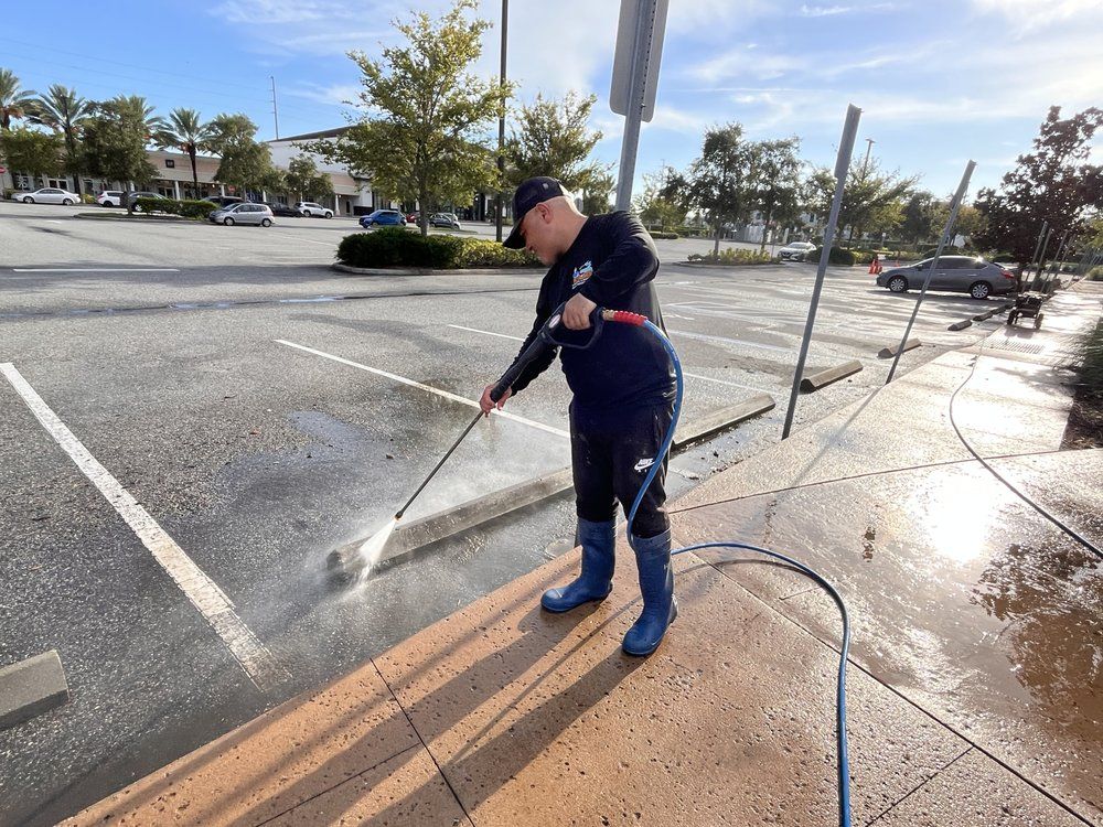 Man power washing a concrete surface in a parking lot. He wears a cap, dark clothing, 