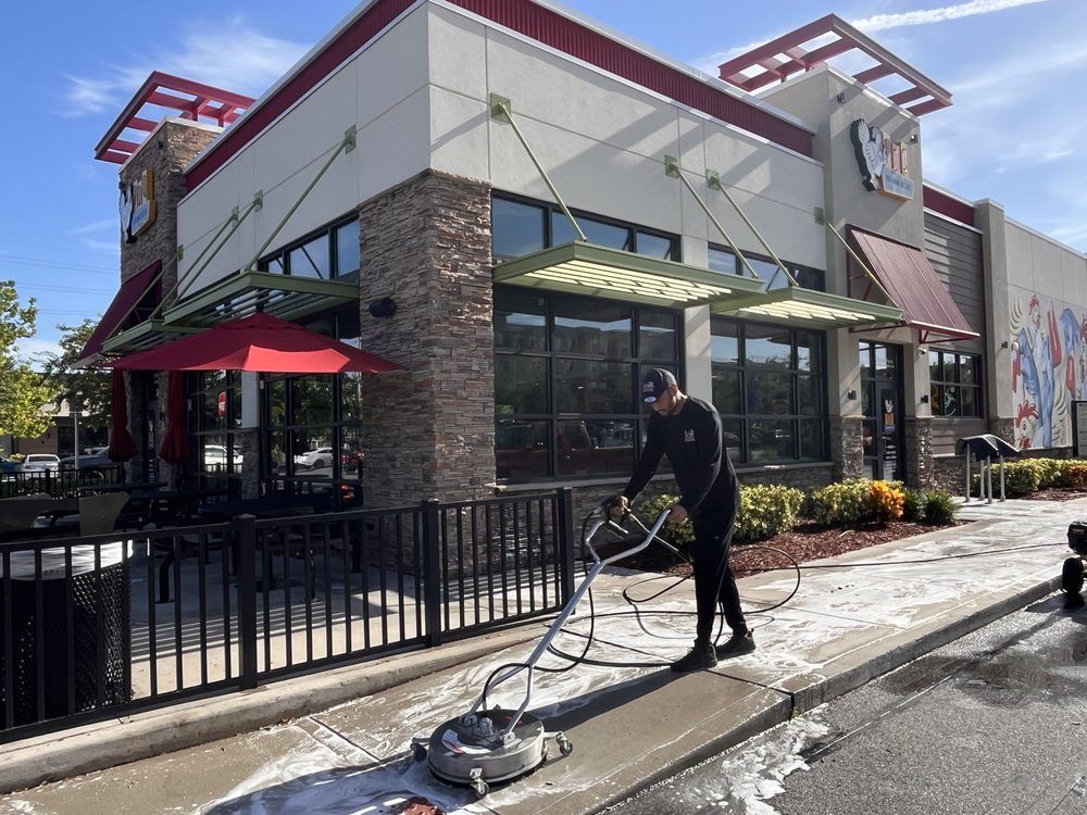 Person power washing a concrete sidewalk in front of a restaurant with a brick and glass facade.