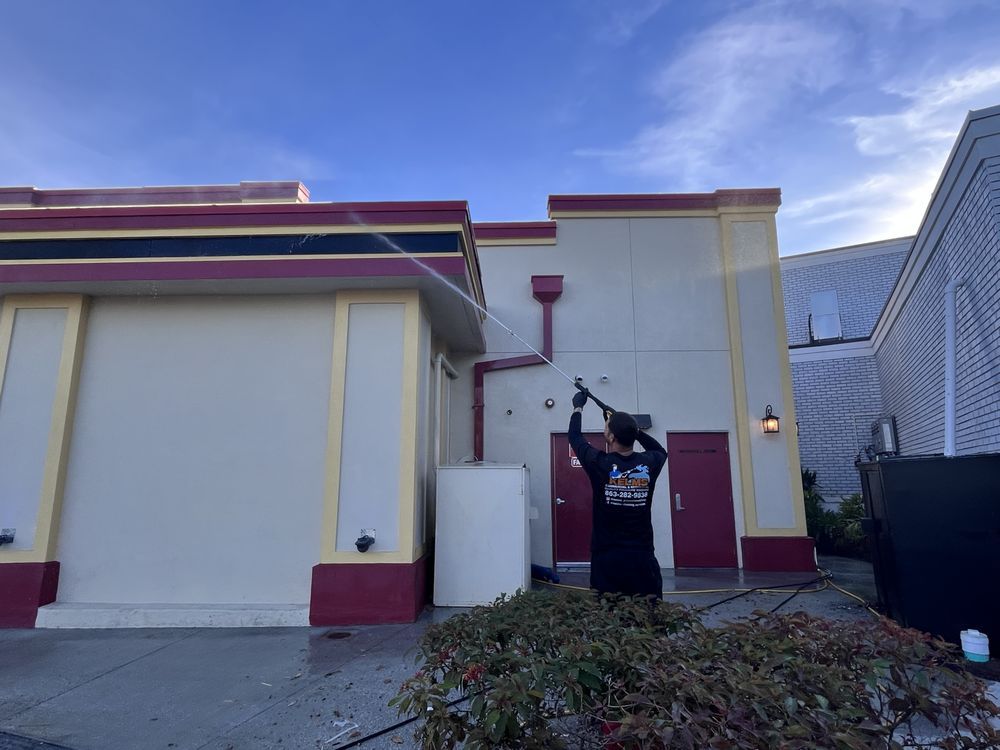 A person power washes a building with beige walls and burgundy trim on a sunny day.