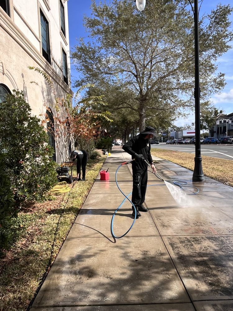 Person pressure washing a sidewalk next to a building on a sunny day. Another person is nearby.