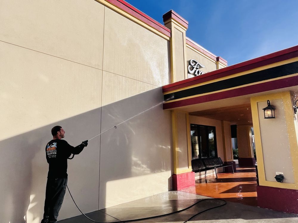 A person pressure washing the exterior of a building with tan walls and a red and black awning on a sunny day.