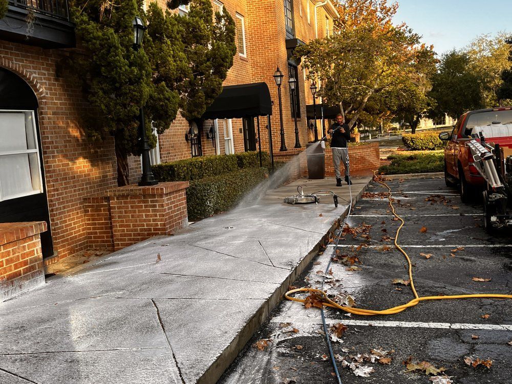 Person spraying a sidewalk with a pressure washer outside a brick building on a sunny day. A red truck is parked nearby.