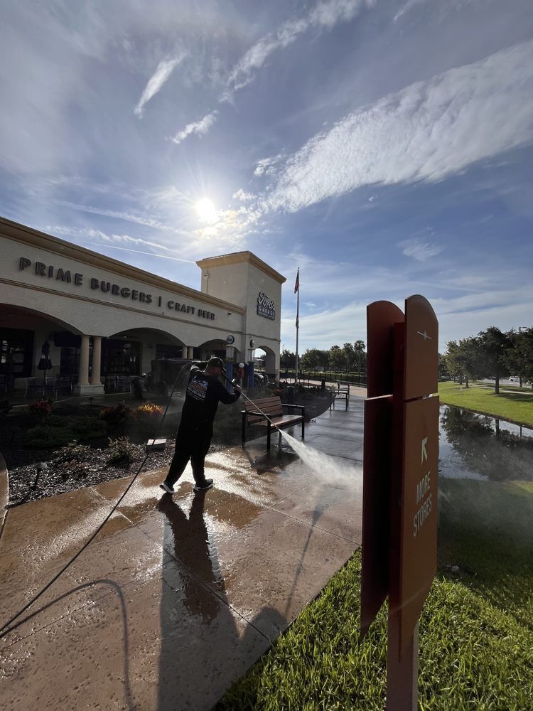 Person pressure washing a walkway outside a building, Prime Burger & Craft Shop, under a bright sunny sky. 