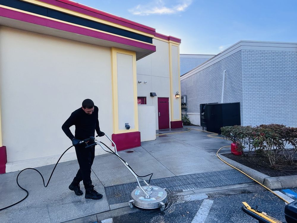 Man pressure washing a concrete surface near a building with red and gold accents. He's wearing all black.