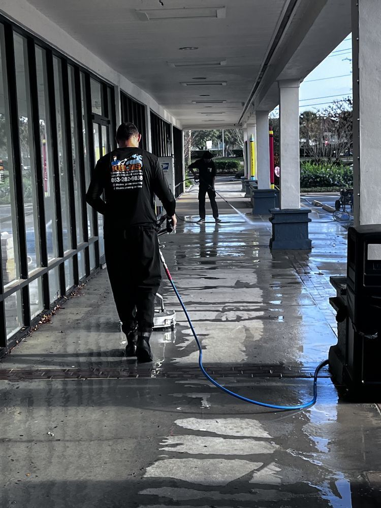 Two workers cleaning a wet concrete walkway outside a store. One pulls a hose, the other is in the background.