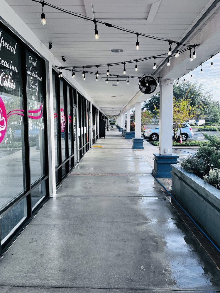 A long, covered walkway with storefronts. String lights hang from the ceiling. The ground is wet.