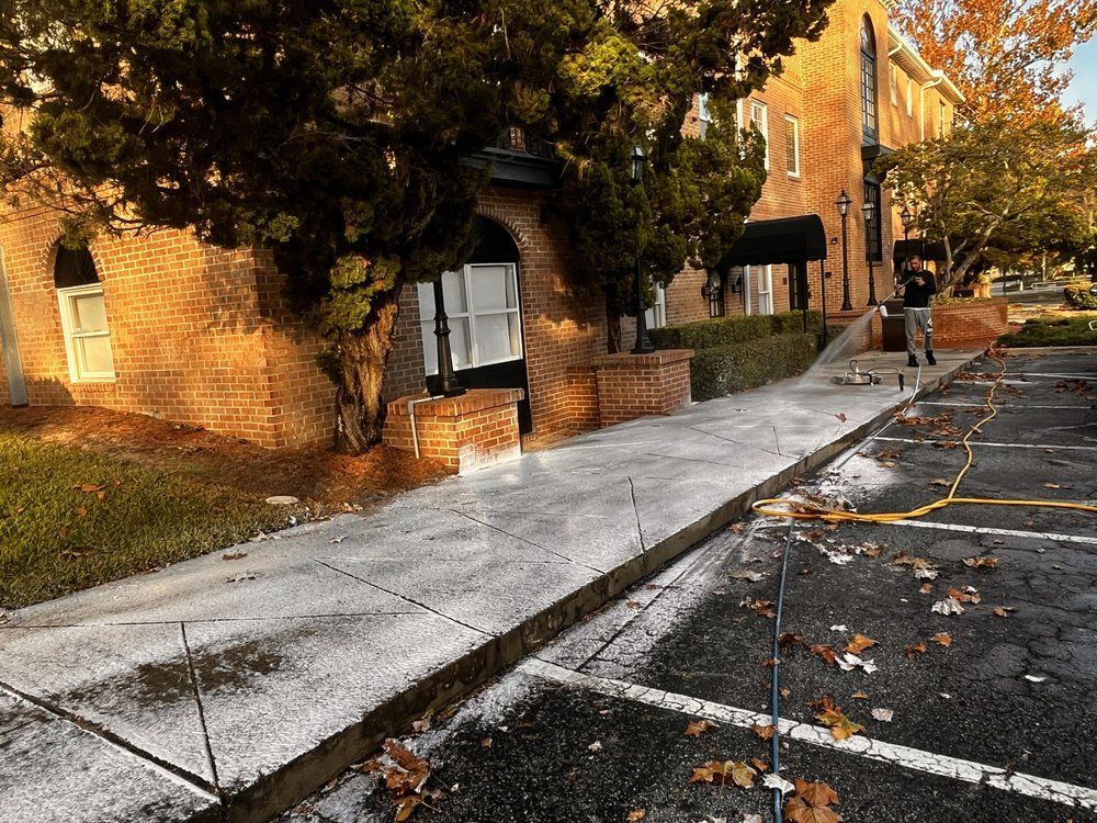 A walkway with snow, bordered by a brick building, an archway, and parking lot with fallen leaves