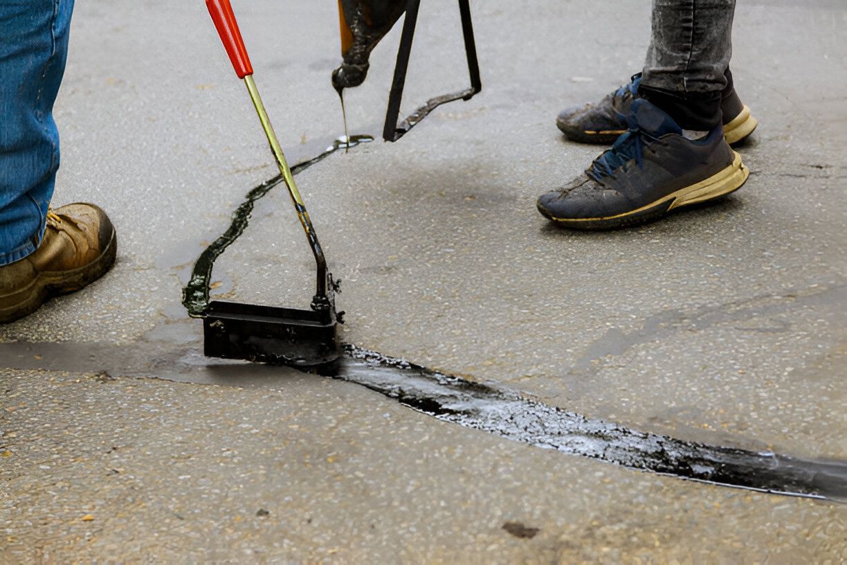 Workers seal a crack in pavement with black sealant and a roller tool.