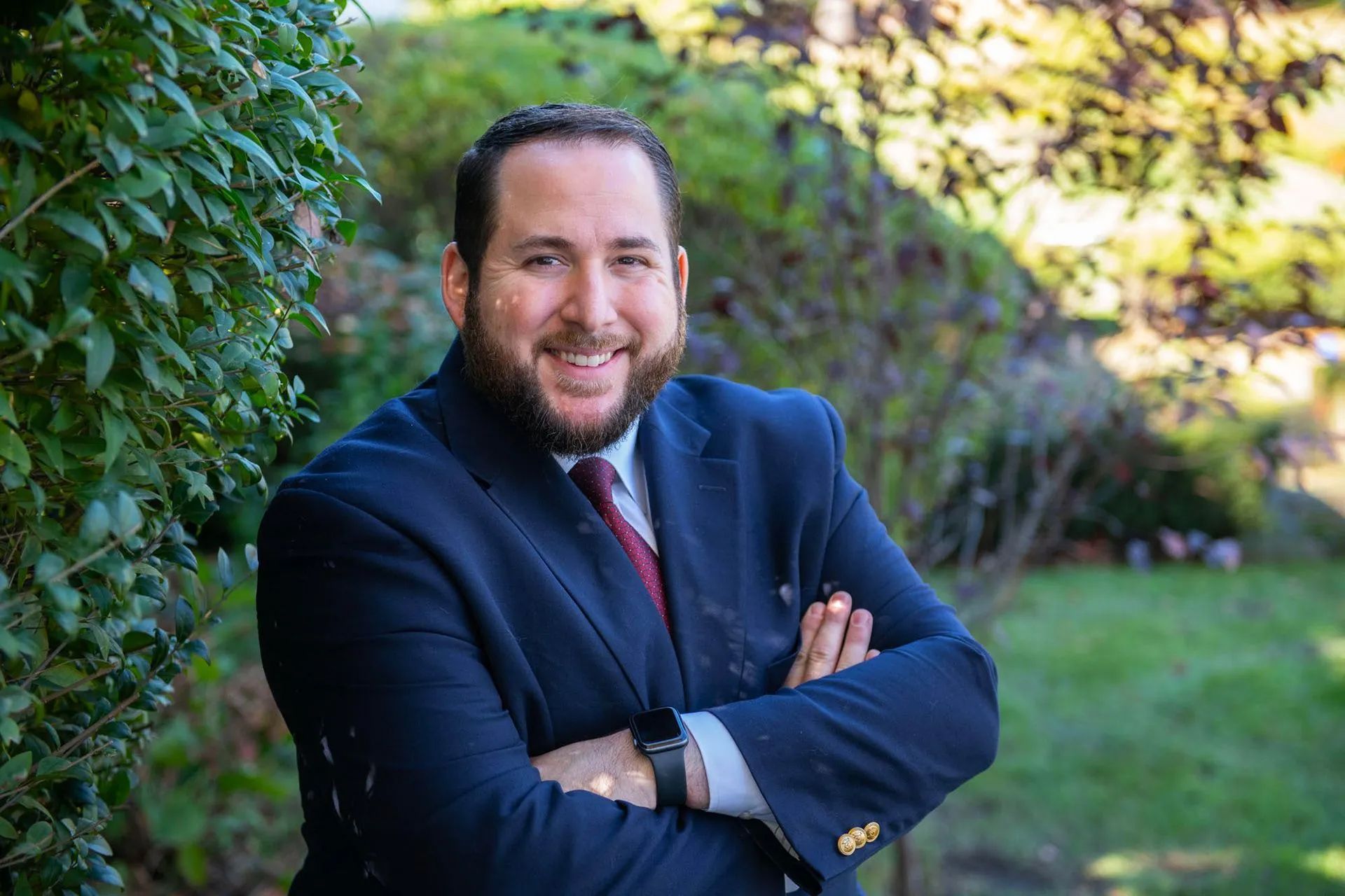 a man in a suit and tie is standing with his arms crossed in front of a bush .