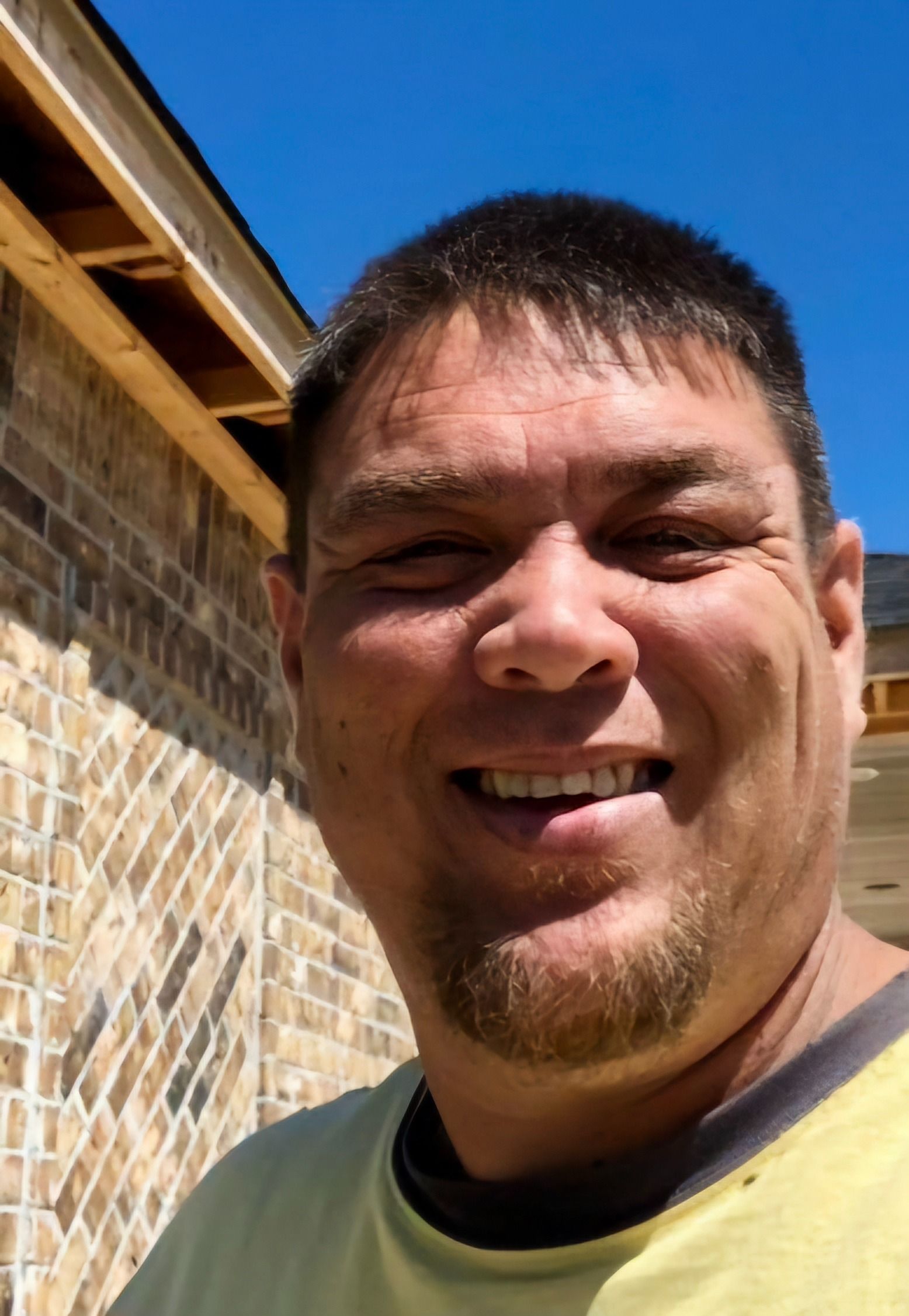 A man in a yellow shirt is smiling in front of a brick wall