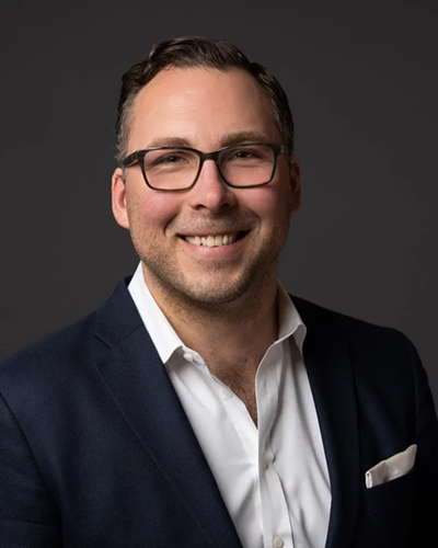 Man in glasses, smiling, wearing a navy blazer and white shirt, with a gray background.