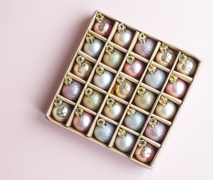 Box of assorted pastel-colored Christmas ornaments with gold caps, arranged on a pink surface.