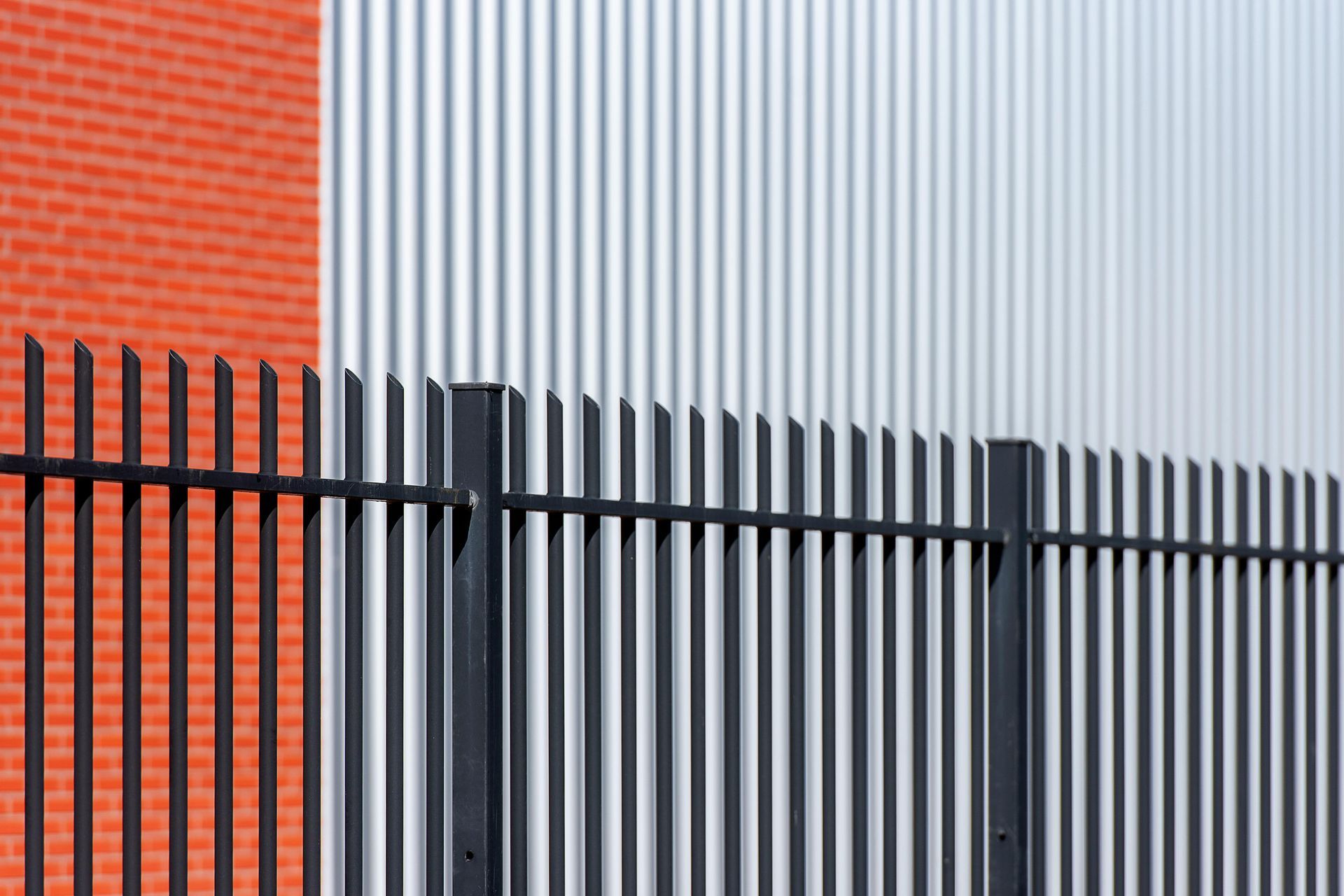 Red brick wall beside silver corrugated metal, with a black metal fence in front.