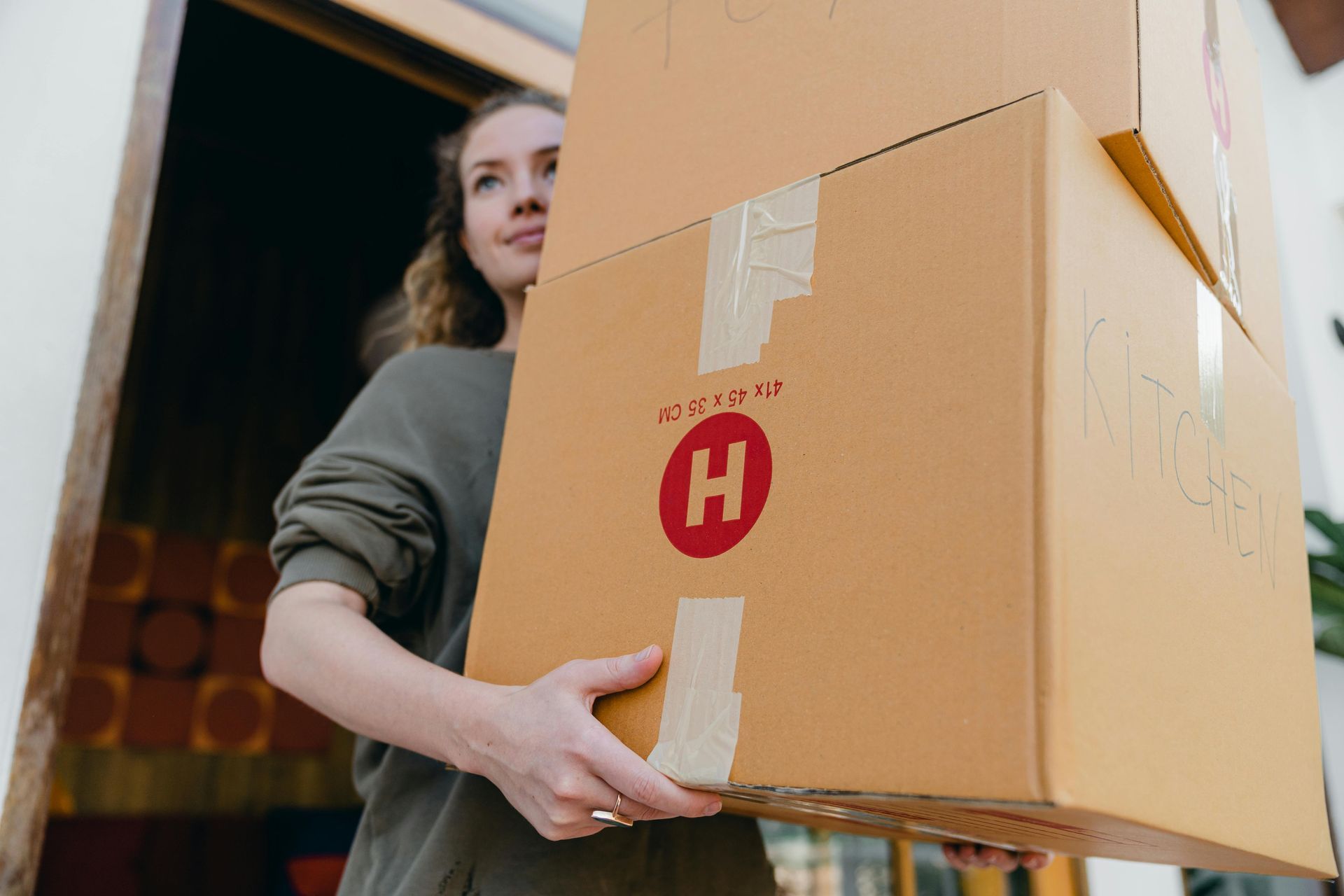Person carrying a large cardboard box indoors, with a red “H” logo visible on the box.
