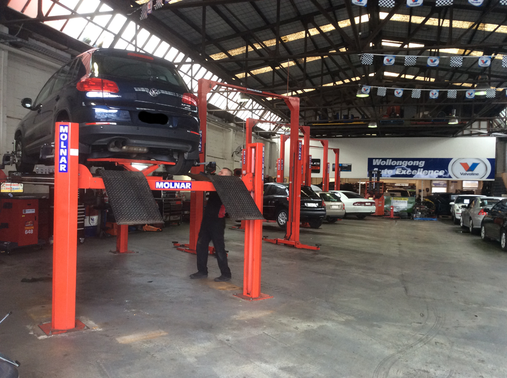 A Car is Sitting on a Lift in a Garage With A Mechanic Fixing It— Wollongong Auto Excellence In Wollongong, NSW