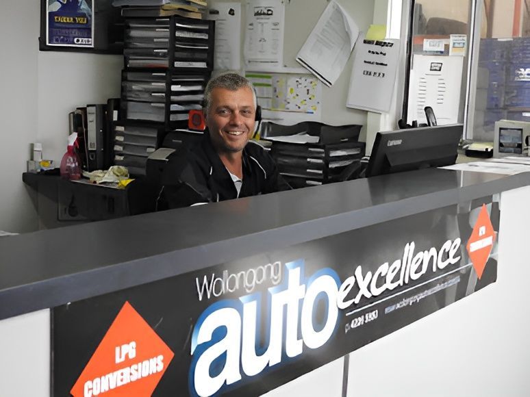 A Man Sitting at a Counter With a Sign That Says Auto Excellence — Wollongong Auto Excellence In Wollongong, NSW