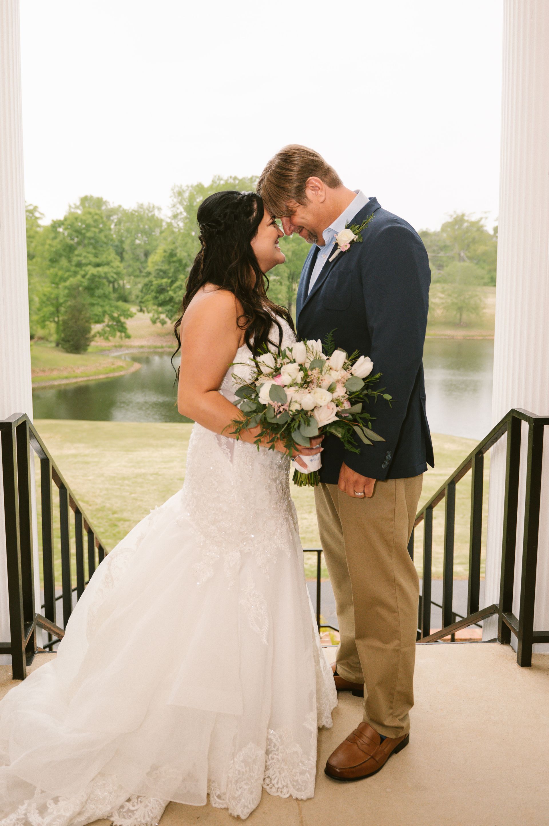 Bride and groom embrace on outdoor steps; she wears a white gown, he wears a navy blazer.