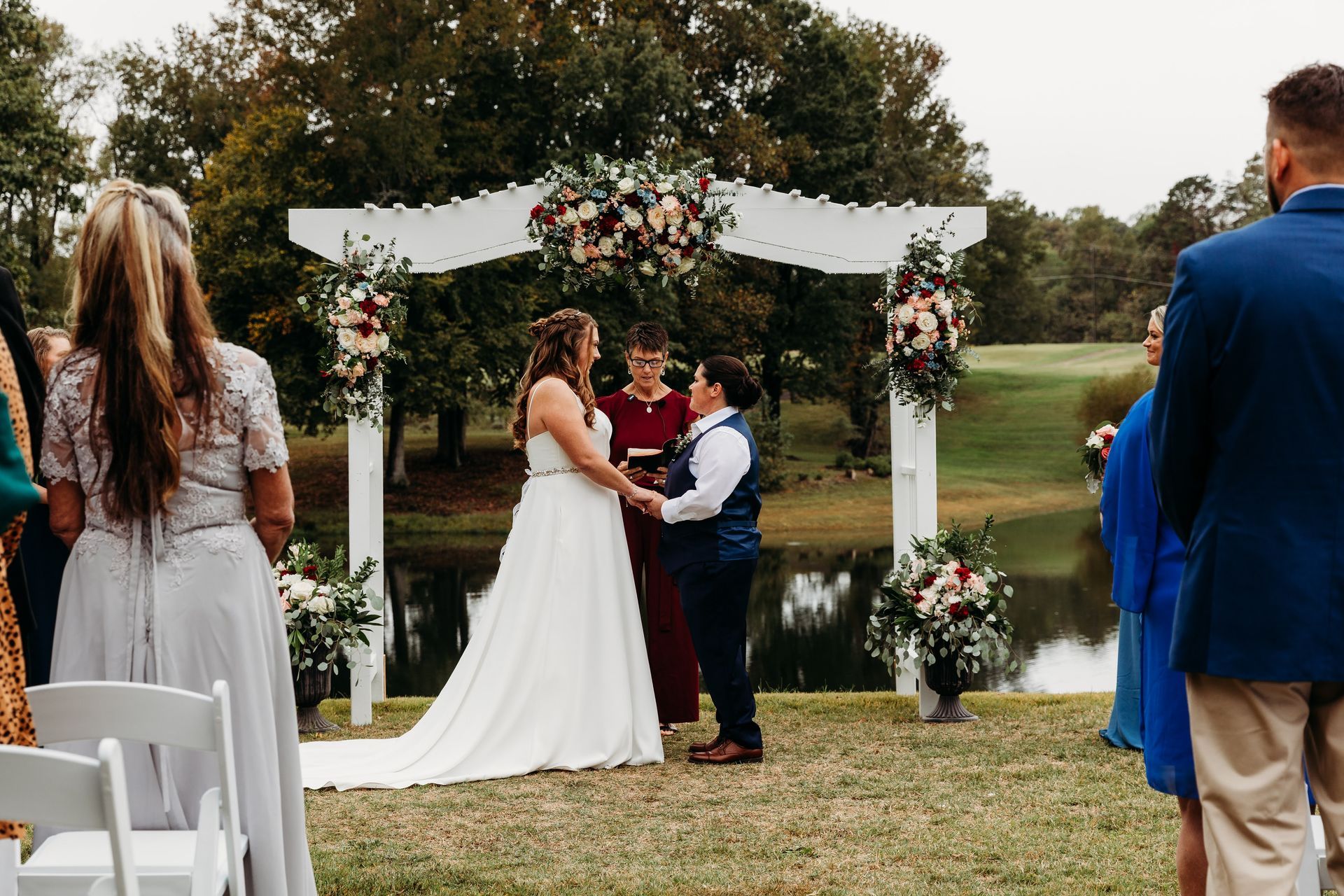 Two people holding hands during an outdoor wedding ceremony, under a floral arch. Guests watch.