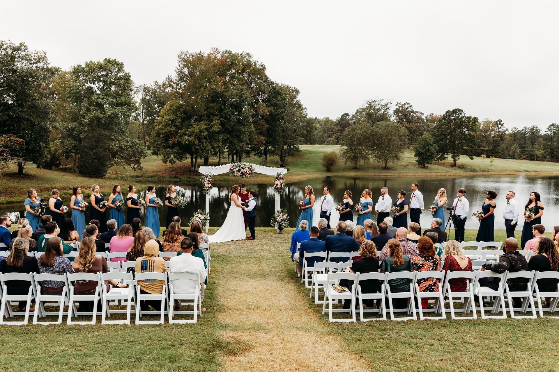 Wedding ceremony by a lake: couple in front, bridesmaids and groomsmen flank them, guests seated on white chairs.